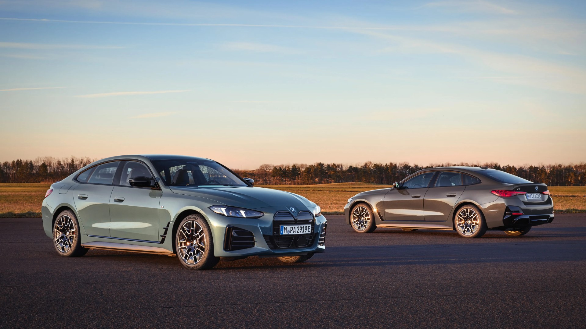 Two BMW electric sedans, one green and one bronze, parked on an asphalt surface with a field and trees in the background.