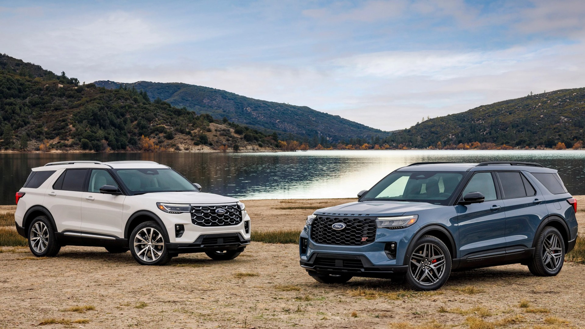 Two Ford Explorer SUVs, one white and one blue, parked on a dirt area near a lake with forested hills in the background.