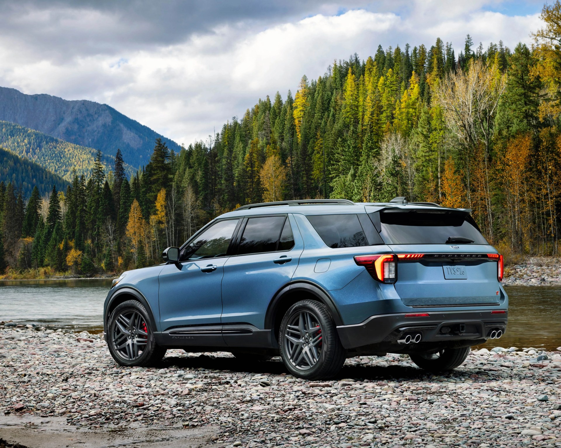 Blue Ford Explorer ST SUV parked on rocky riverbank with forest and mountains in the background.