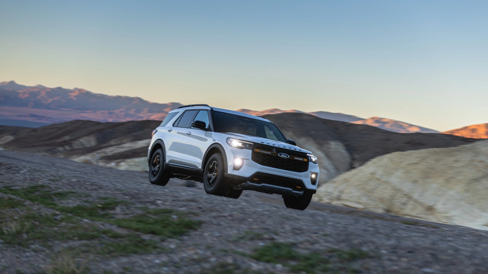 White Ford Explorer SUV driving on a rocky dirt road with mountains in the background at sunset.