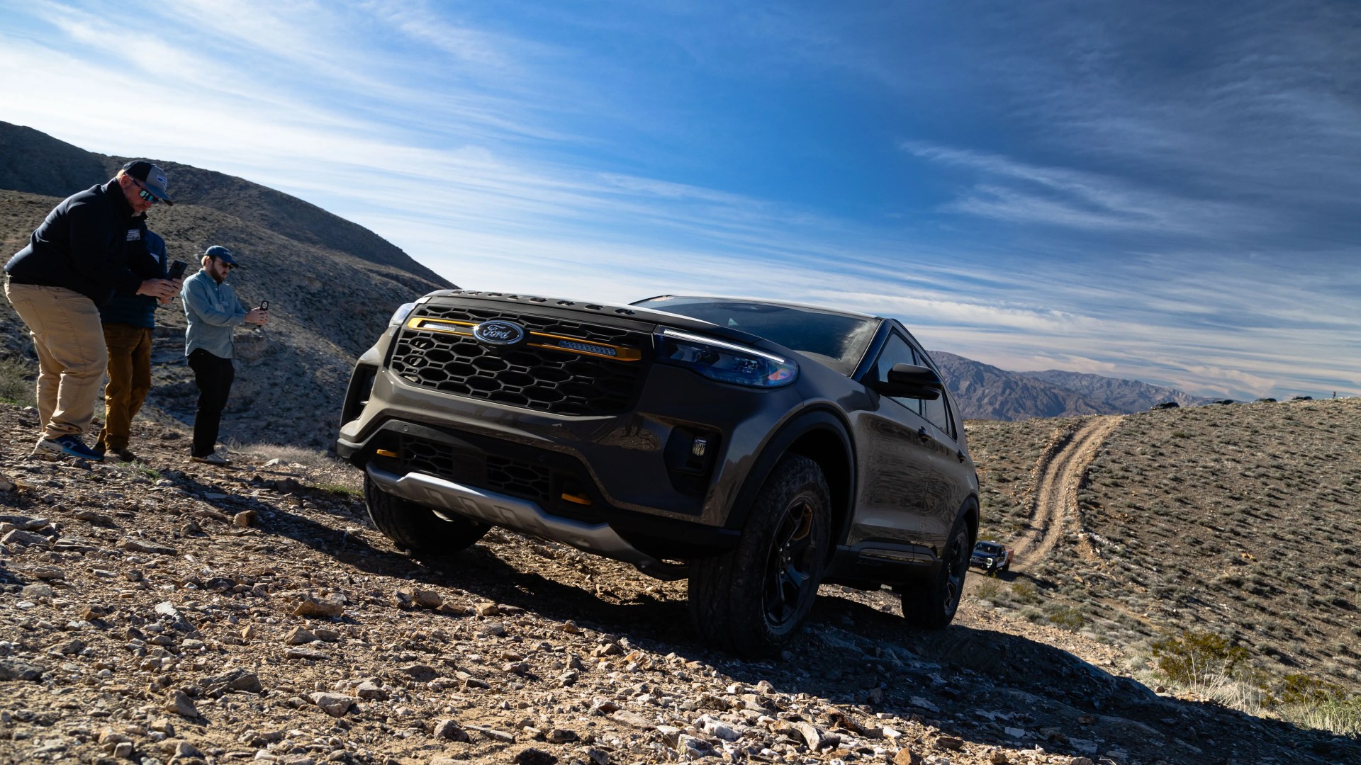 Dark gray Ford SUV driving on rocky off-road terrain with three people photographing it nearby.