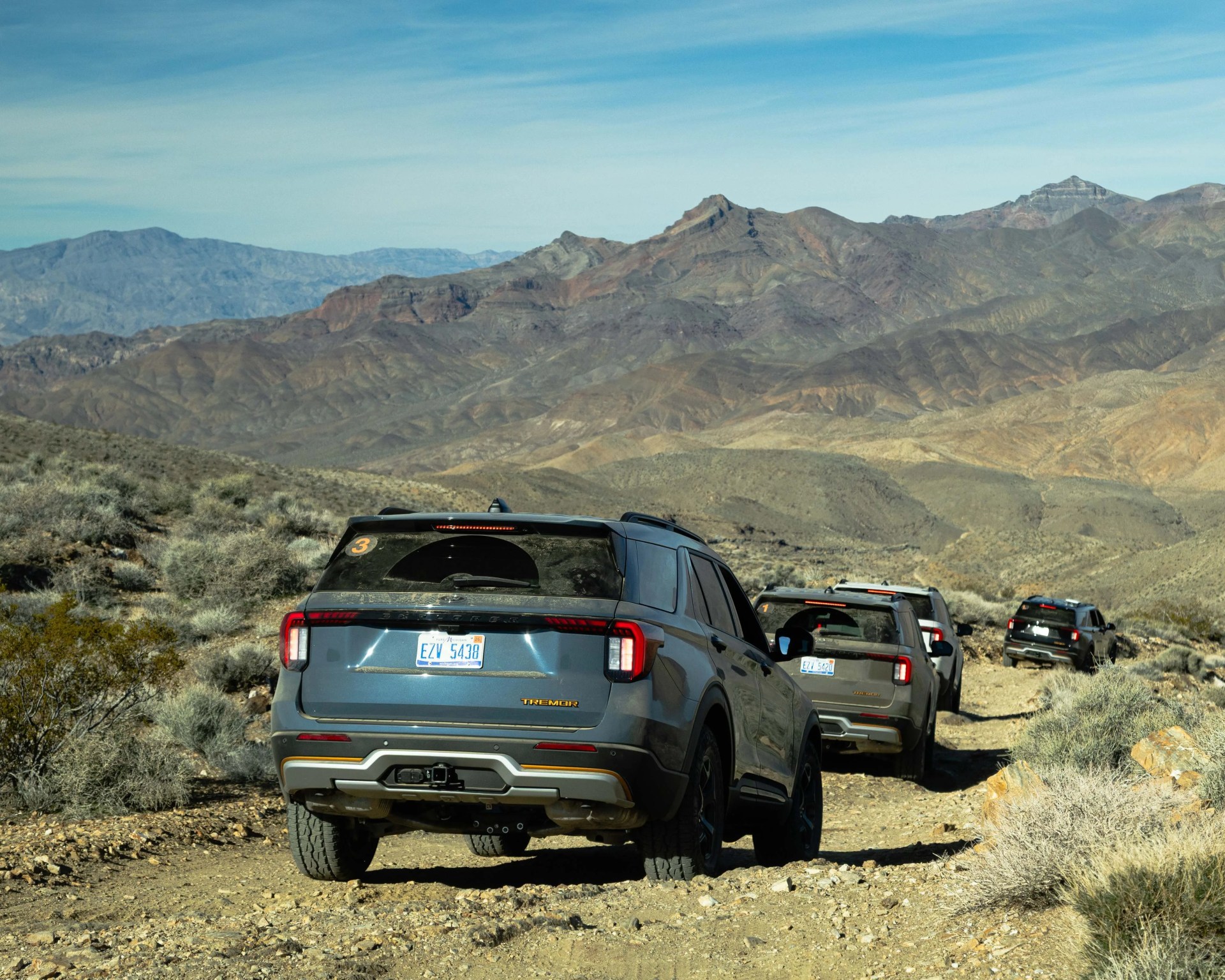 Four gray Ford Bronco SUVs driving on a rocky desert trail with mountains in the background.
