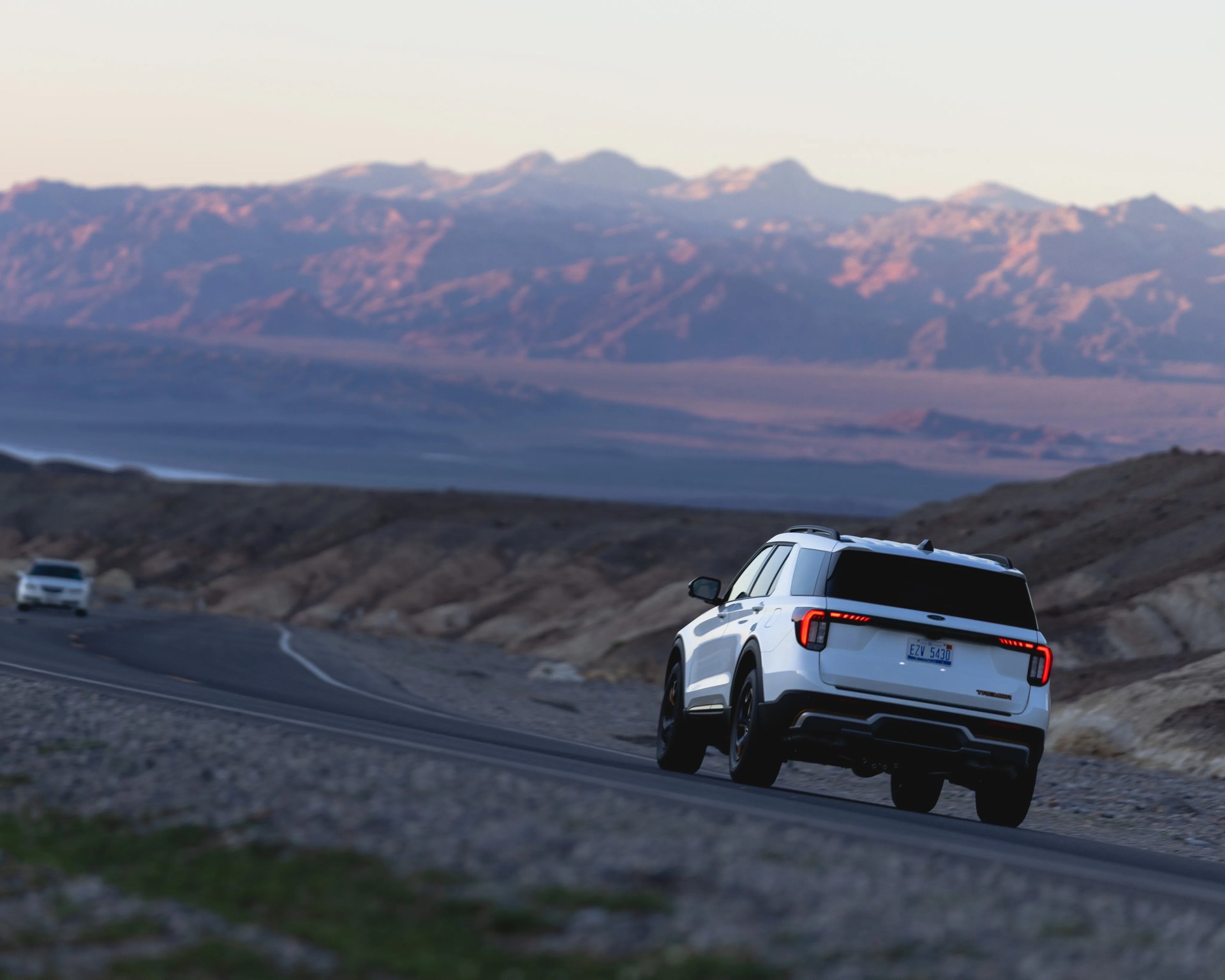 White SUV with black trim driving on a winding road in a mountainous desert landscape.