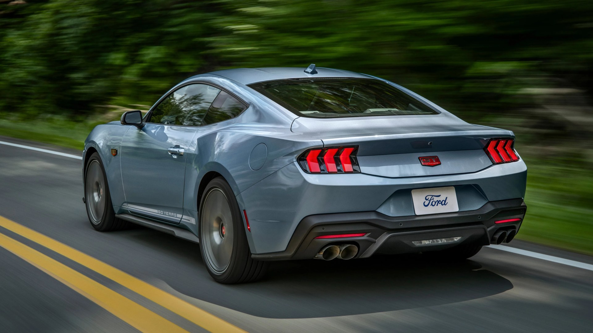 Gray Ford Mustang GT with 60 Years badge driving on a road with blurred green trees in the background.