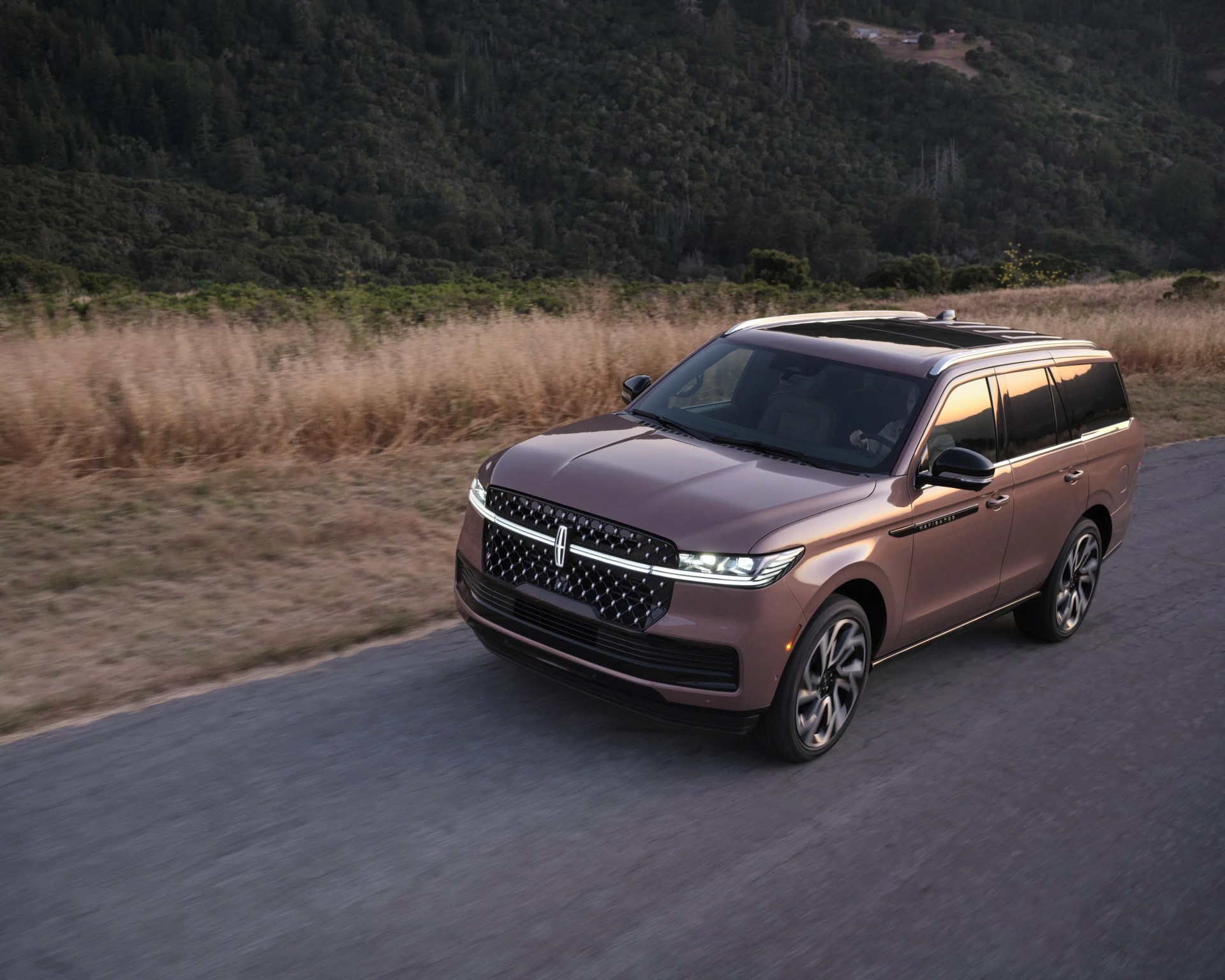 Metallic brown Lincoln Navigator SUV driving on a rural road with forested hills in the background.