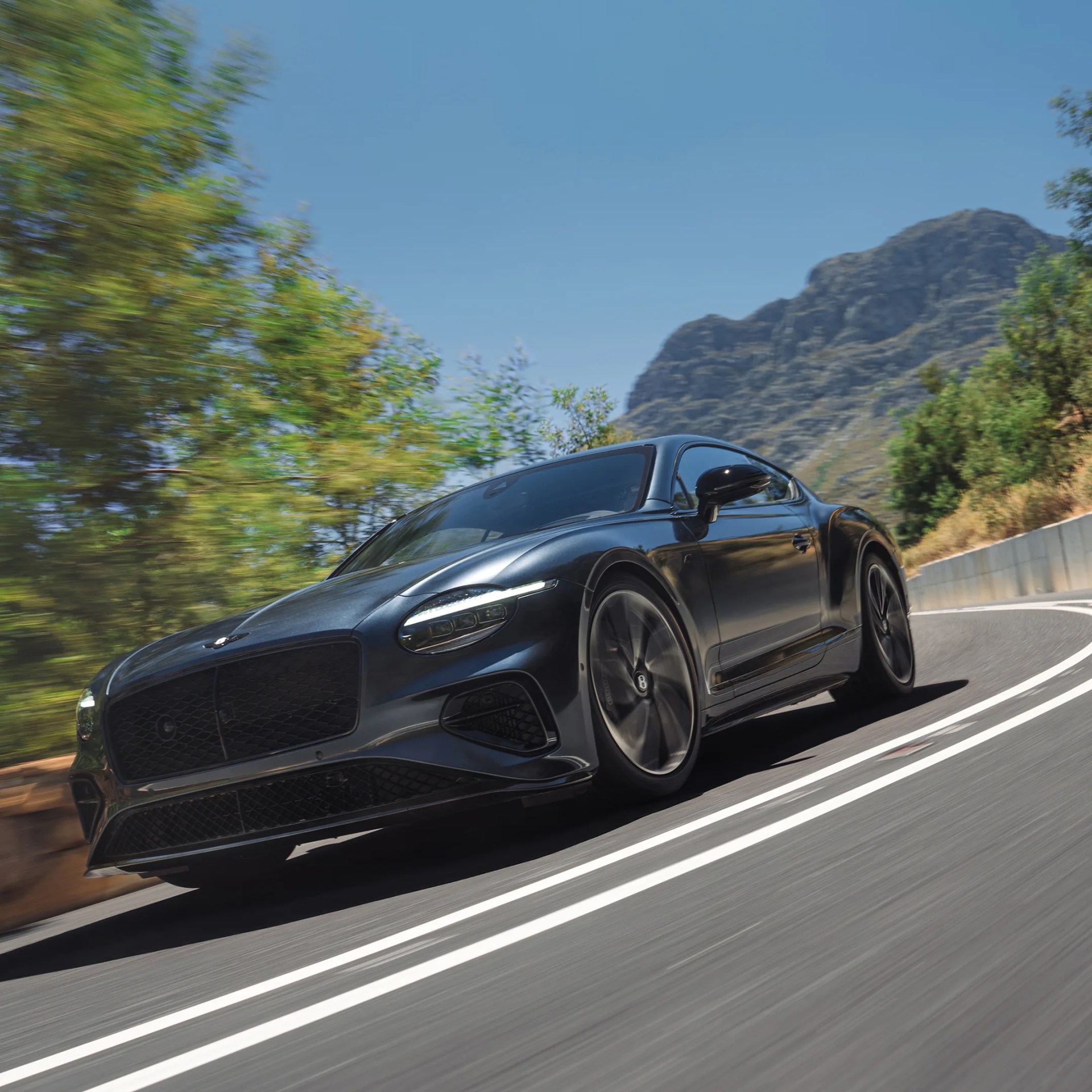 Black Bentley luxury coupe driving on a winding mountain road with blurred trees and rocky hills.