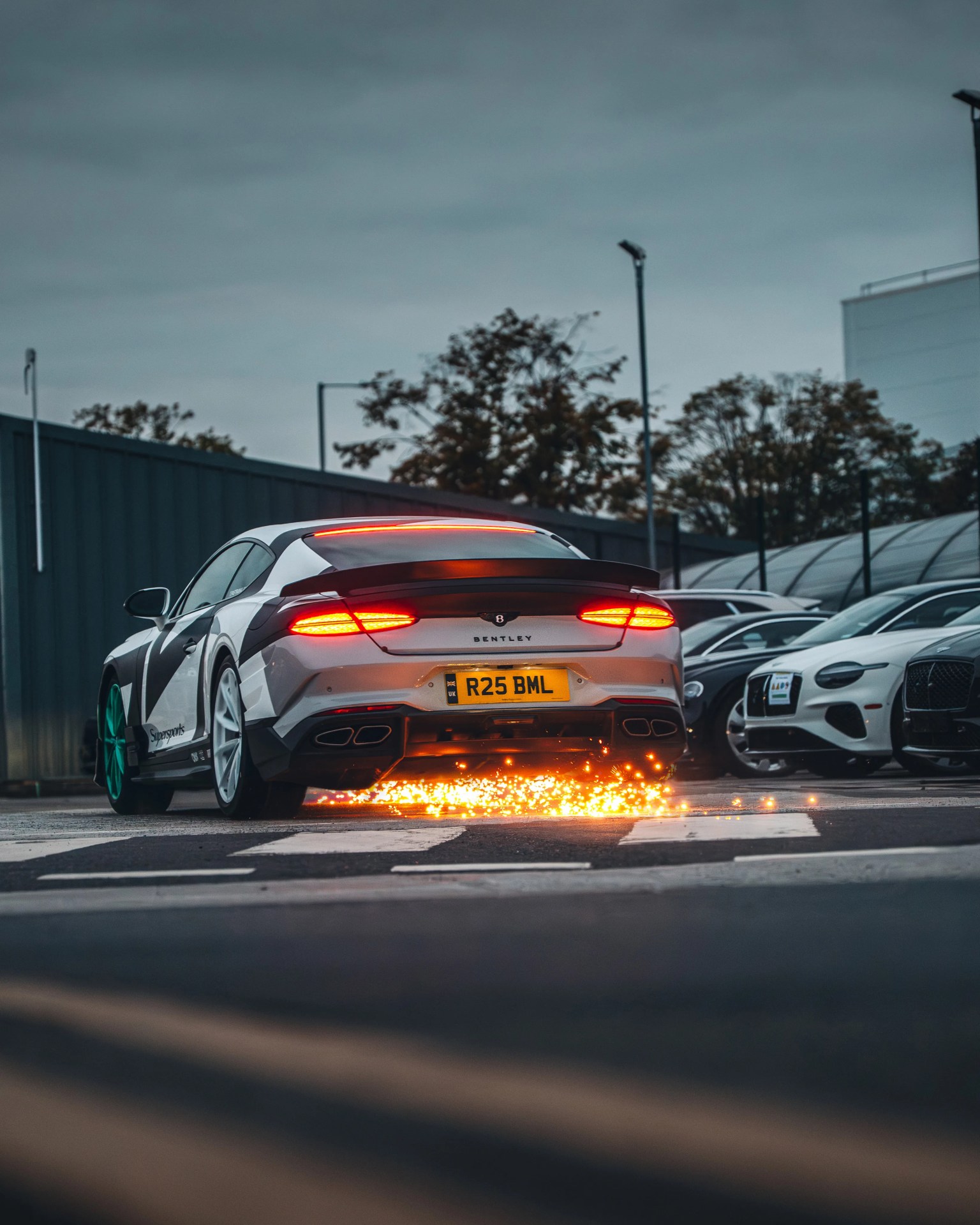 White and black Bentley sports car with green front wheels sparking underneath on a street at dusk.