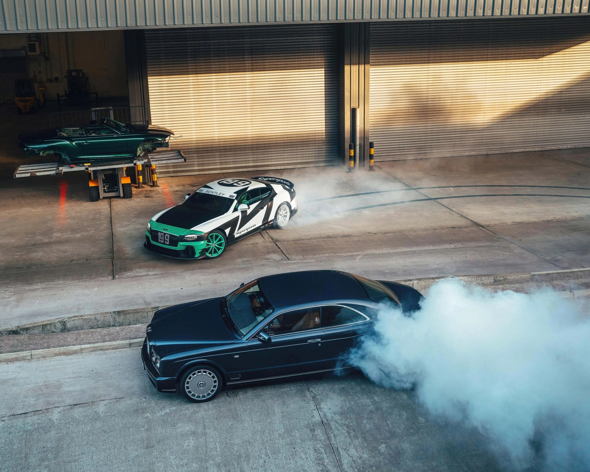 Two luxury cars, one black emitting thick white smoke from the rear tires, and one white and green with racing decals, parked on concrete near a garage door.