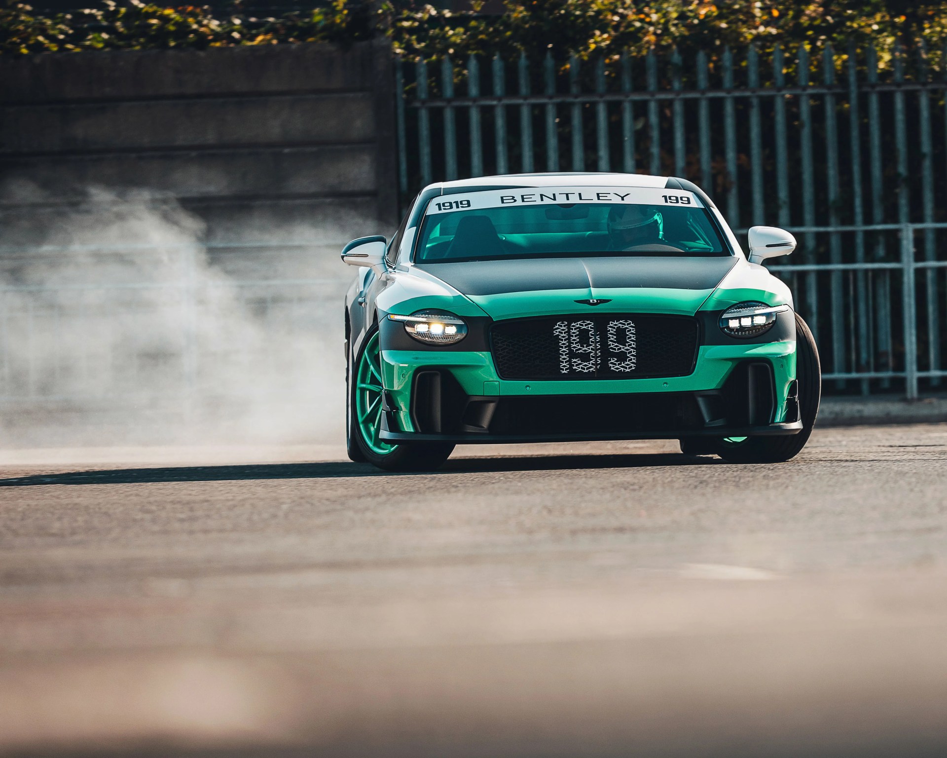 Green and black Bentley sports car drifting on pavement with smoke behind it.