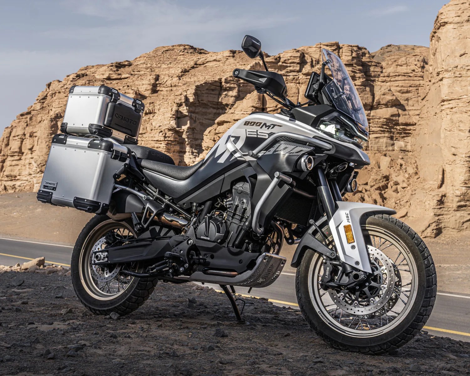 Silver and black CFMOTO 800MT adventure motorcycle with aluminum panniers parked on rocky terrain near desert cliffs.