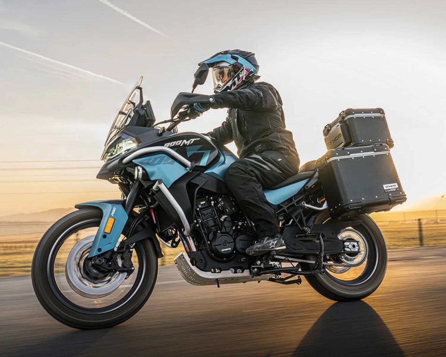 Blue and black CFMOTO 800MT adventure motorcycle with rider in black gear on a road at sunset.