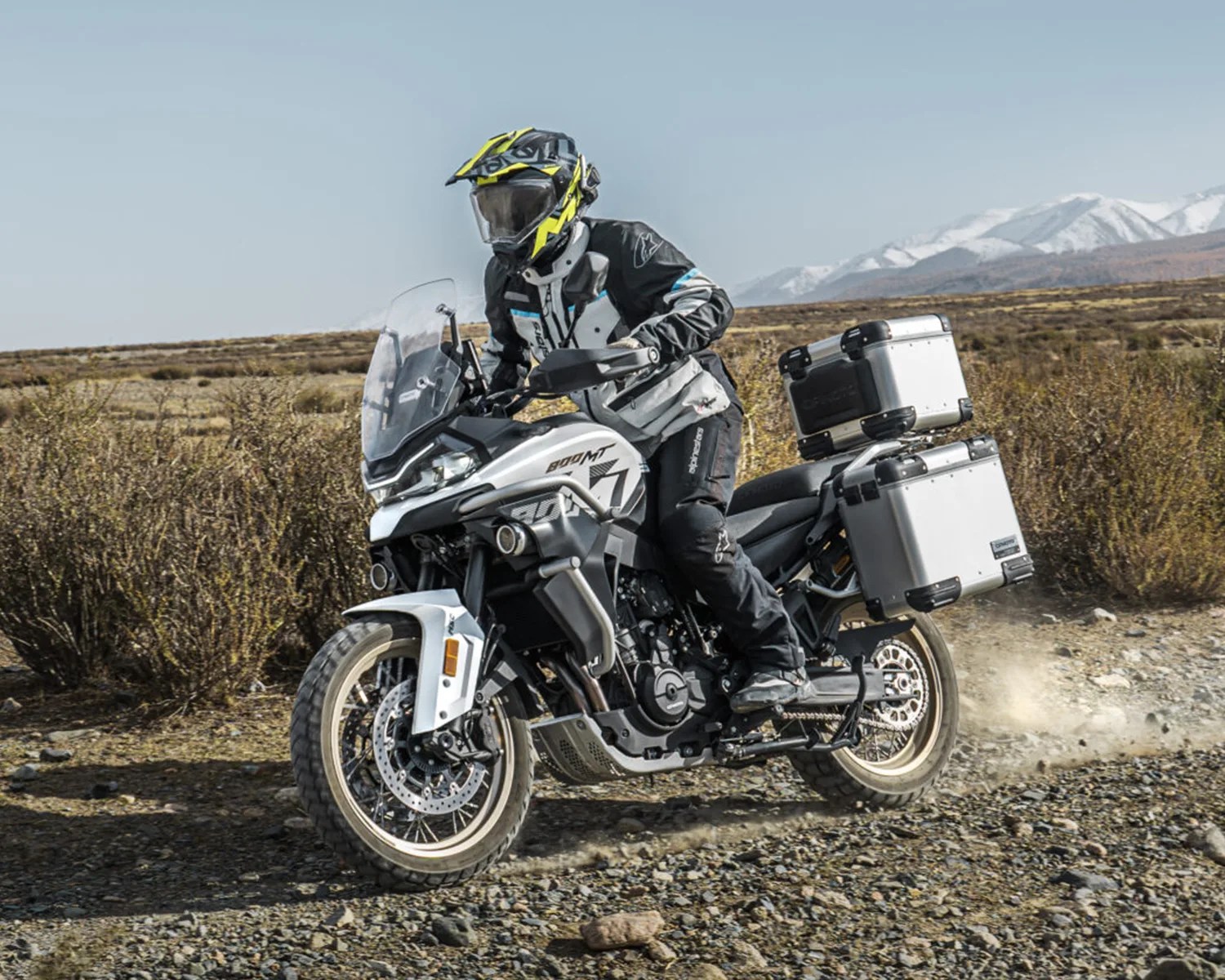 Motorcyclist wearing black and white gear with yellow helmet riding a white adventure motorcycle on a rocky trail with mountains in the background.
