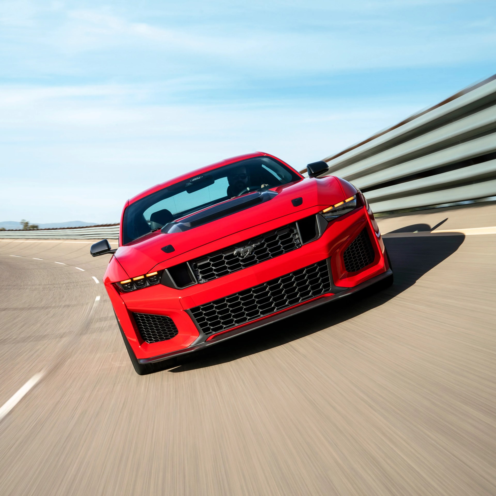Red Ford Mustang with black hood scoop driving on a curved racetrack with metal guardrails.