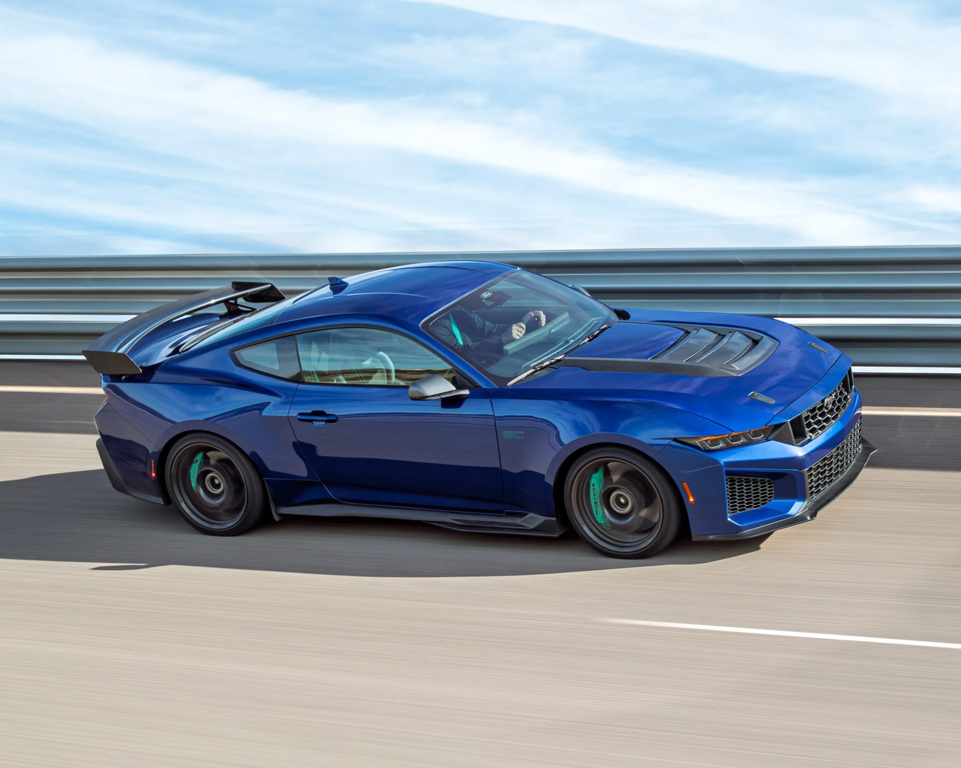 Blue sports car with black hood vents and rear spoiler driving on a highway with metal guardrails.
