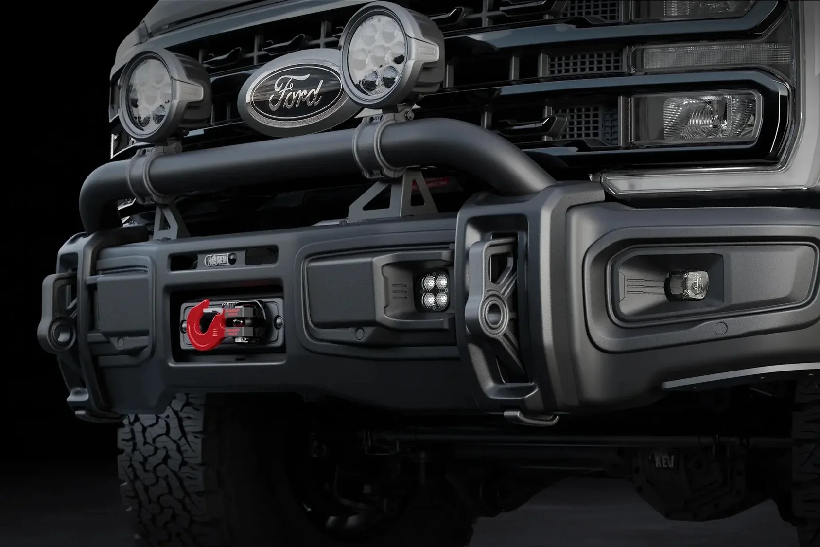 Close-up of a black Ford truck front bumper with off-road lights, a red tow hook, and rugged tires.