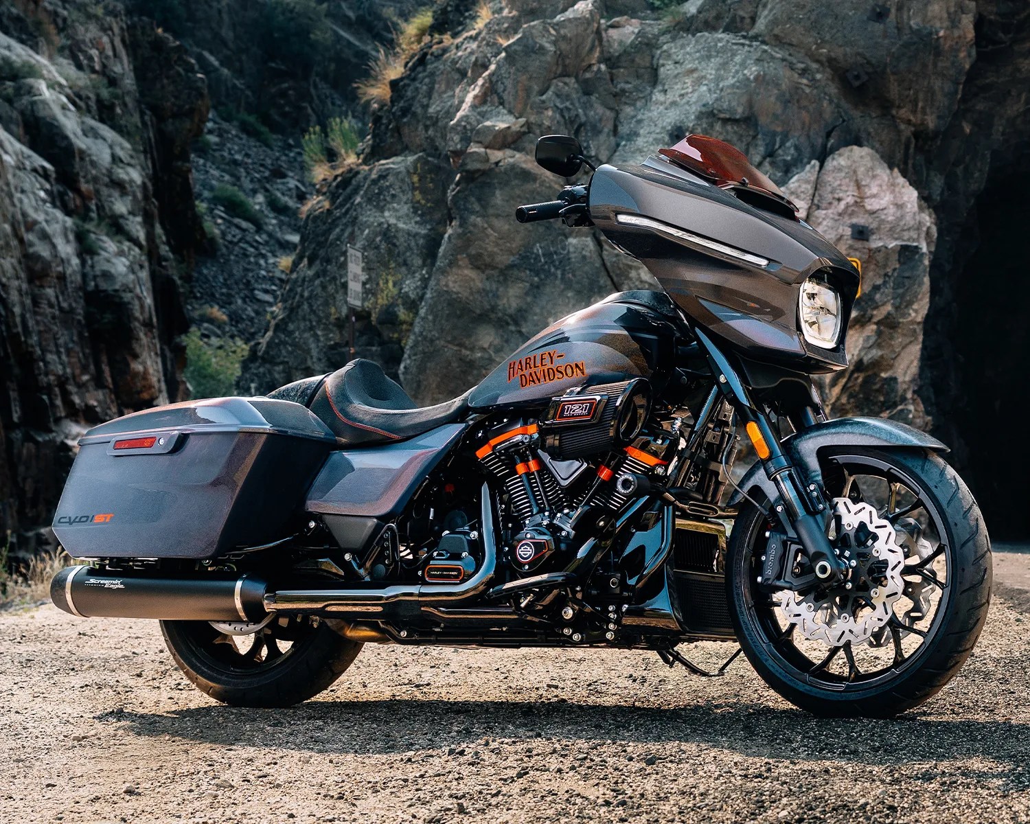 Matte gray Harley-Davidson touring motorcycle with saddlebags parked on a gravel road in front of rocky cliffs.