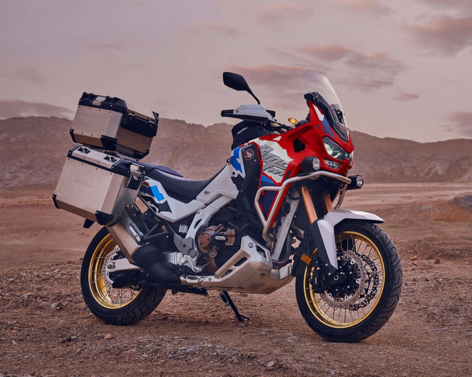 Adventure motorcycle with red, white, and blue bodywork, gold spoked wheels, and aluminum panniers on a dirt road.