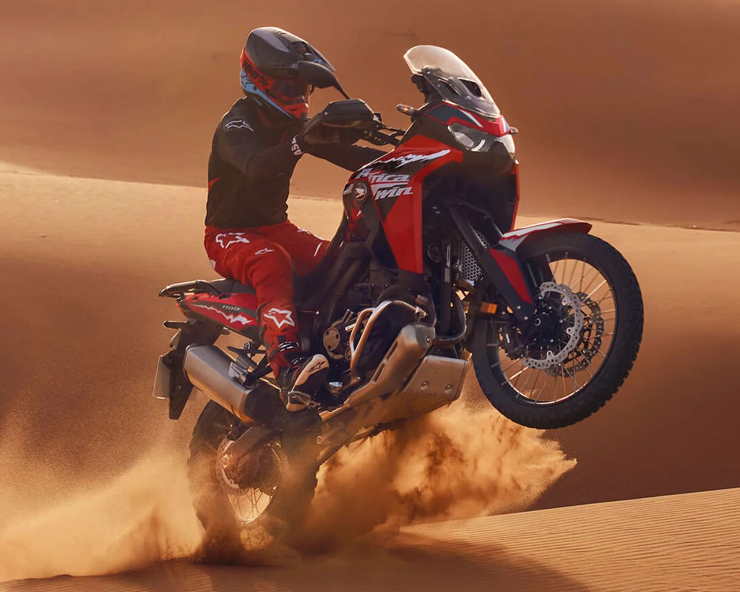 Red and black adventure motorcycle performing a wheelie on sand dunes with rider in red and black gear.