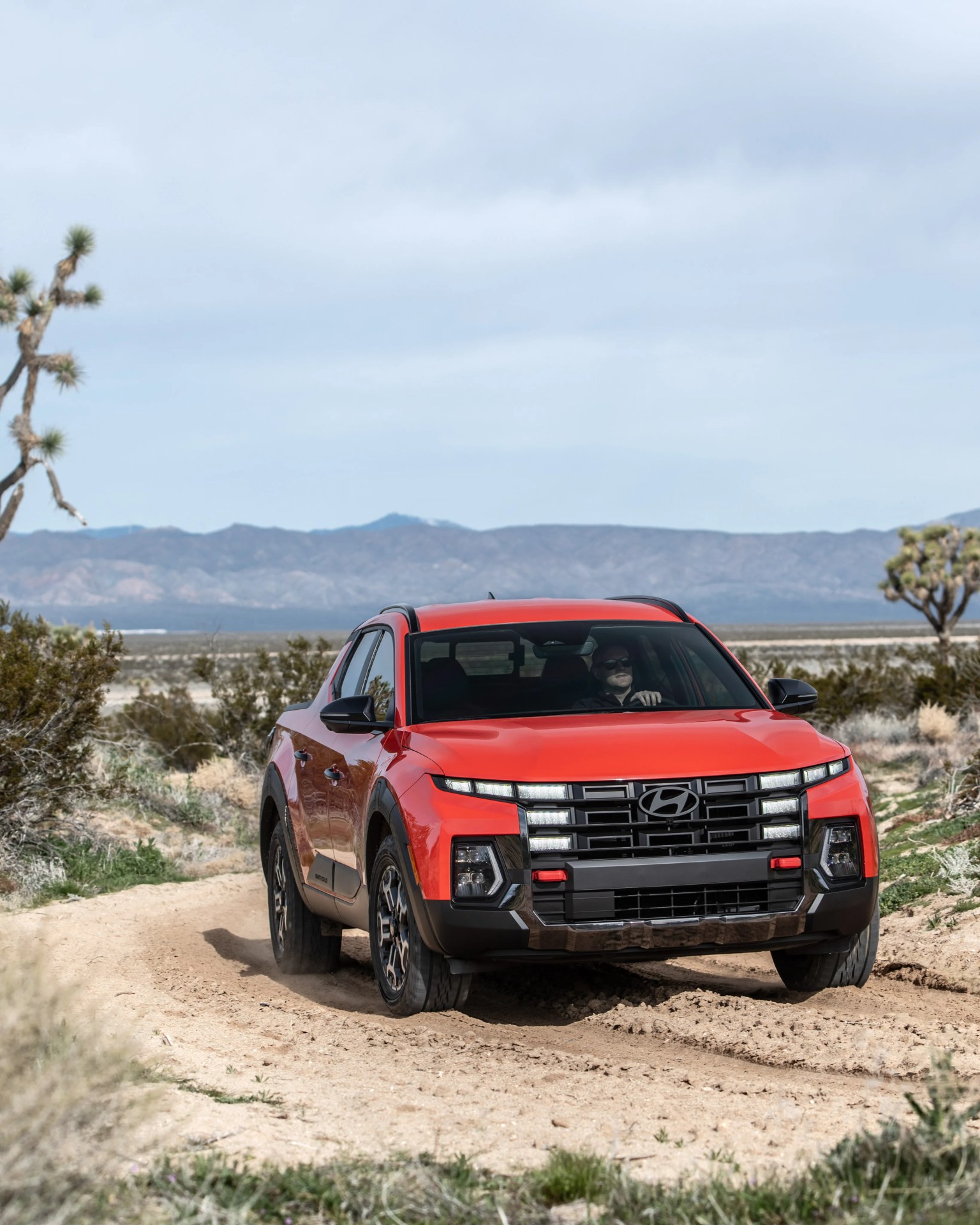 Red Hyundai pickup truck driving on a dirt road in a desert landscape with mountains in the background.