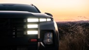 Front close-up of a black vehicle with illuminated LED headlights at sunset in a natural setting.