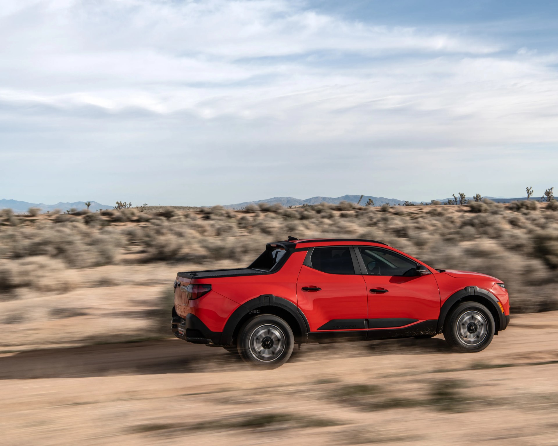 Red four-door pickup truck driving on a dirt road in a desert landscape with mountains in the background.