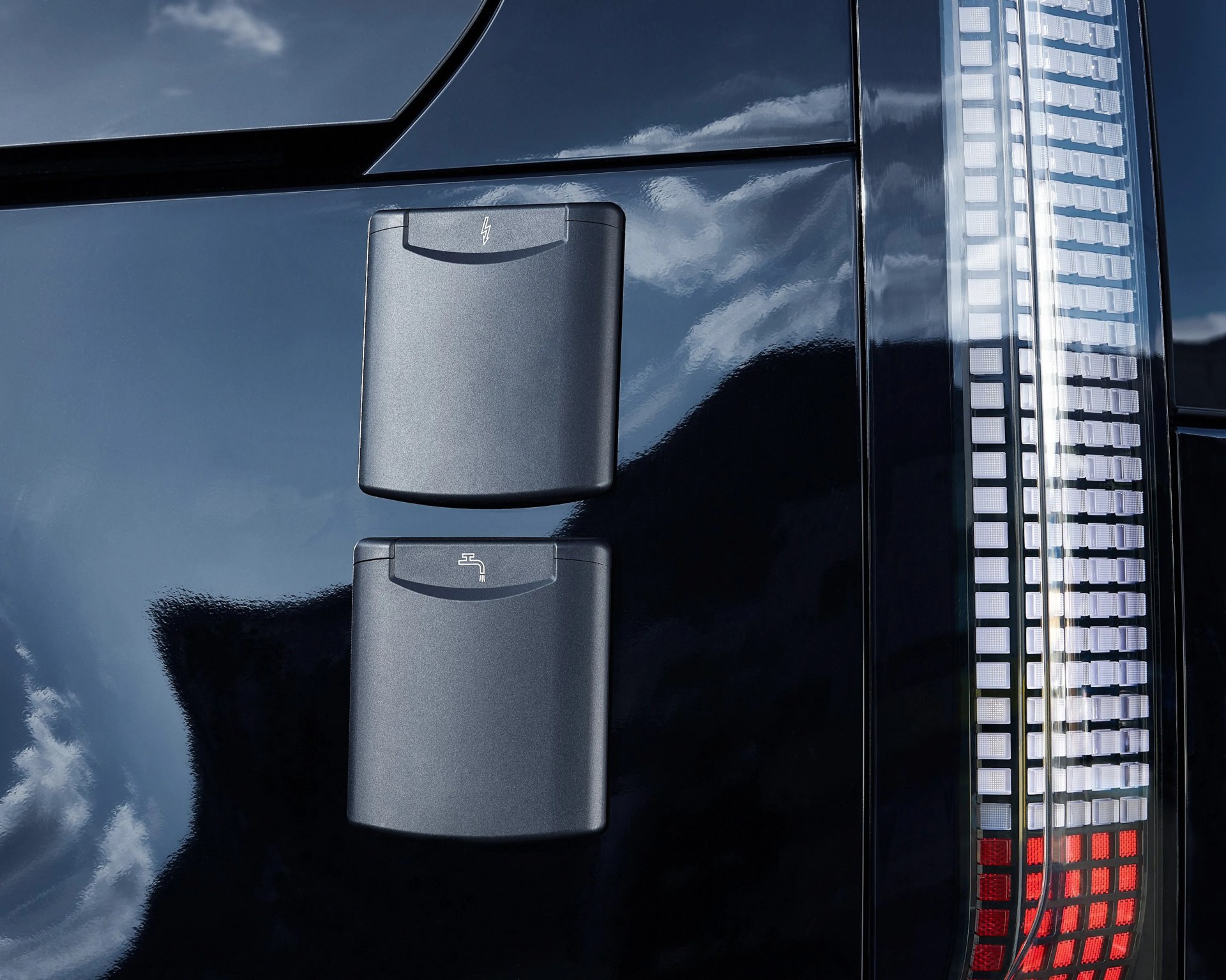 Two black rectangular ports with electric and water icons on the side of a glossy dark vehicle near a vertical taillight.