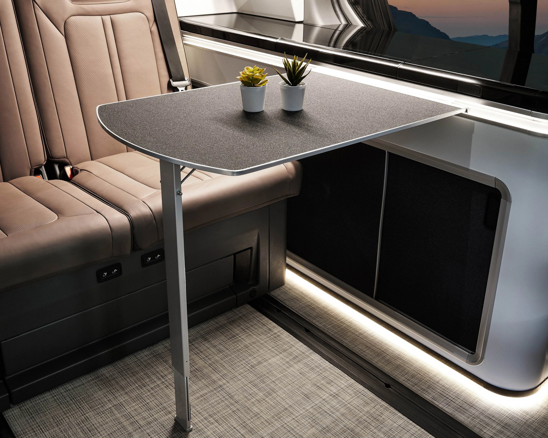Rectangular black foldable table with silver edges and a single leg, attached inside a vehicle next to beige leather seats.