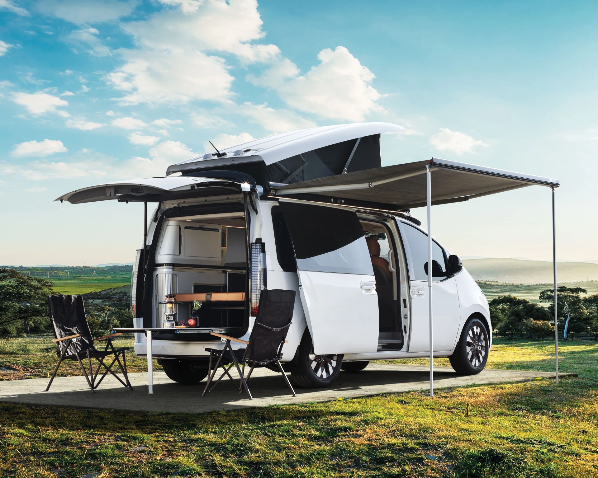 White camper van with pop-up roof and extended awning parked on grass with two black folding chairs and a small table outside.