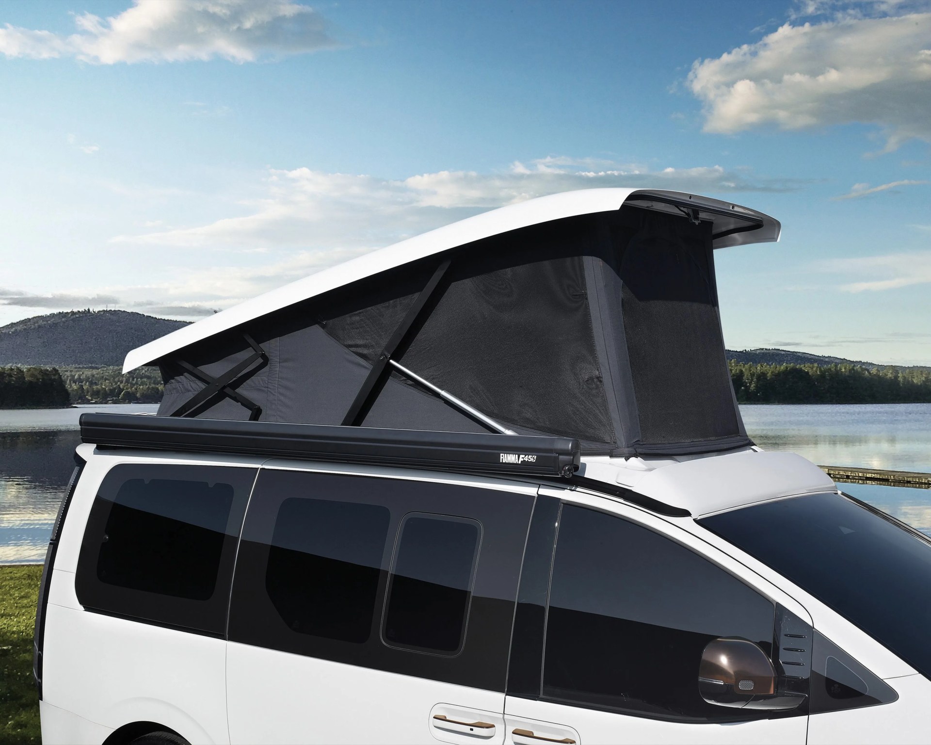 White camper van with black pop-up roof tent parked near a lake under a partly cloudy sky.