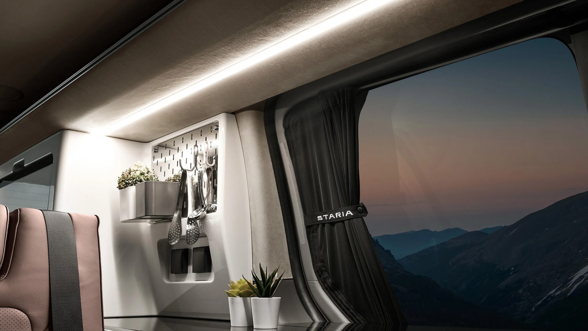 Interior corner of a vehicle with kitchen utensils hanging on a white panel, potted plants, and a window with black curtains labeled "Staria.