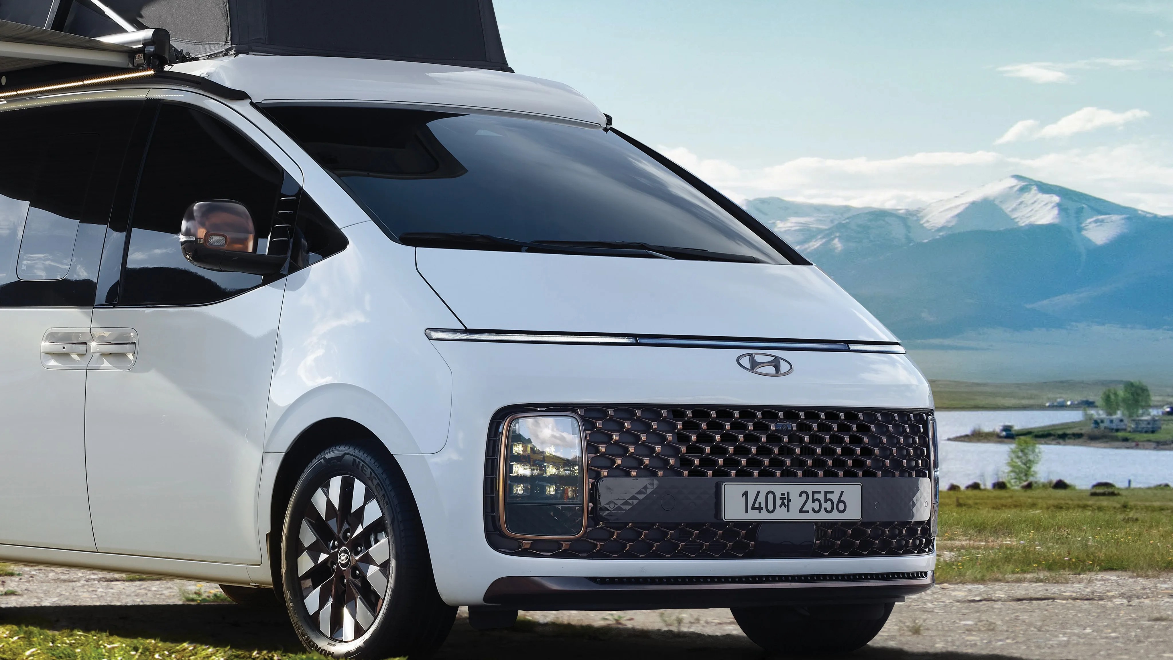 White Hyundai van with black honeycomb grille and roof tent parked on grass with mountains in background.