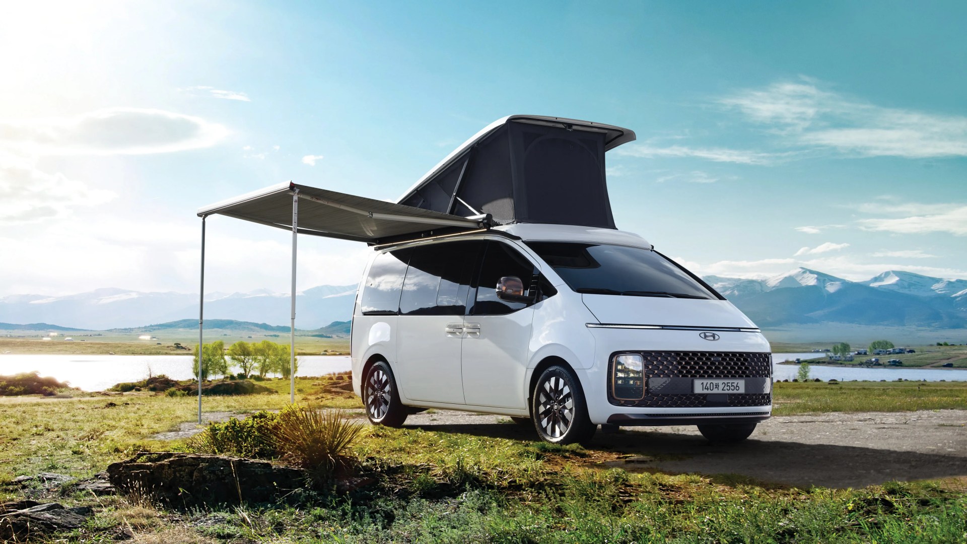 White Hyundai camper van with pop-up roof and extended side awning parked on grassy field near a lake.