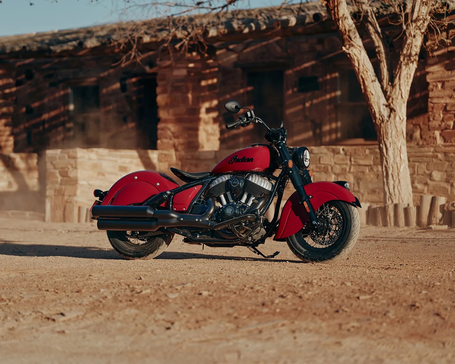 Red Indian motorcycle with black exhaust pipes parked on a dirt ground in front of a stone building.