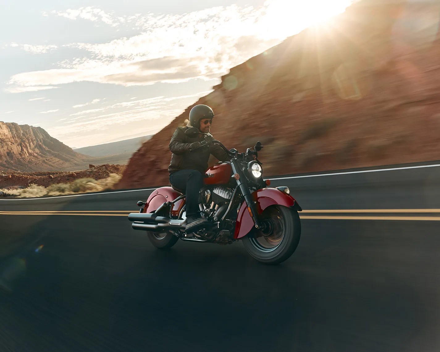 Person wearing a black helmet and jacket riding a red motorcycle on a desert road with rocky hills in the background.