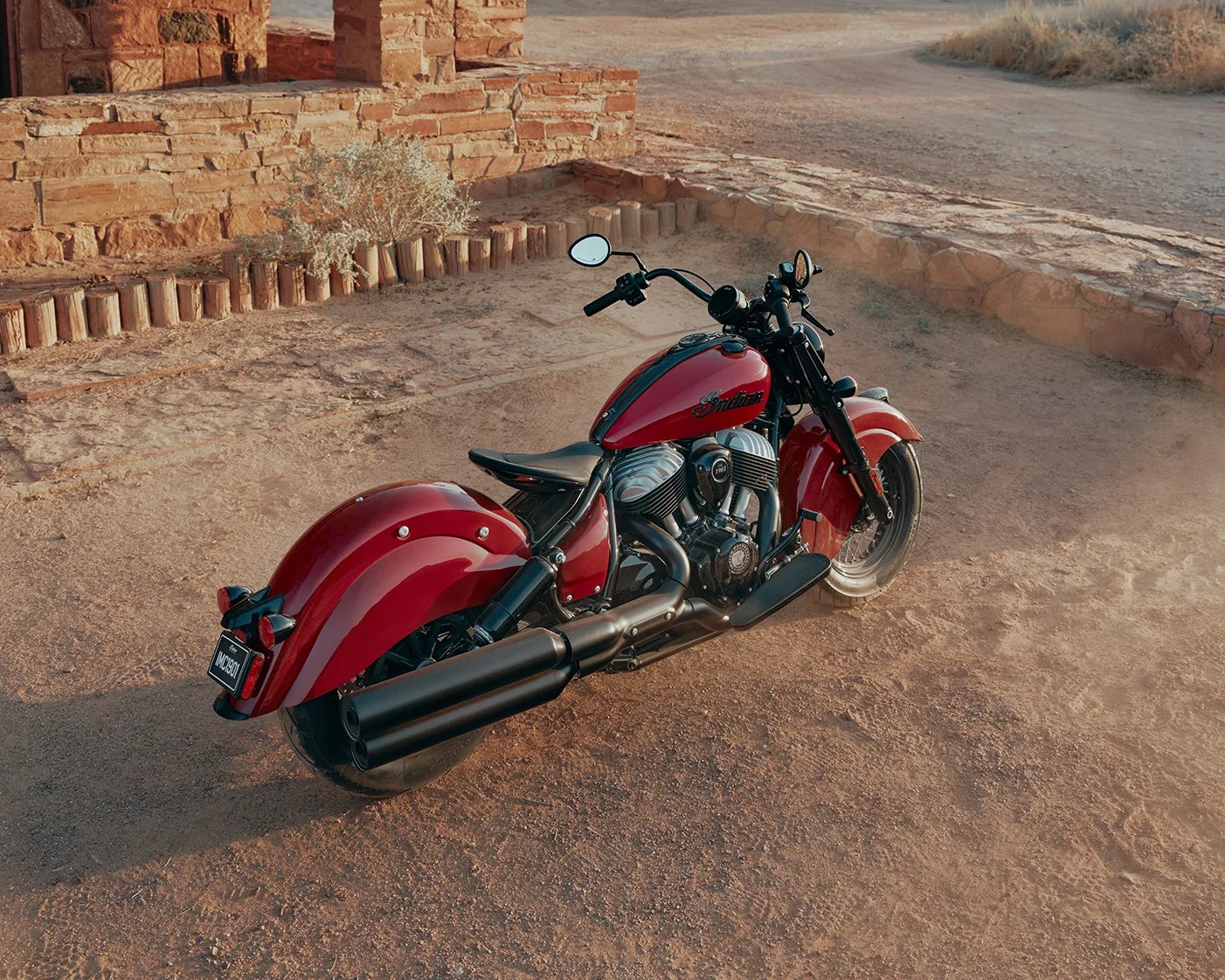 Red Indian motorcycle with black exhaust pipes parked on a dirt surface near a stone wall.