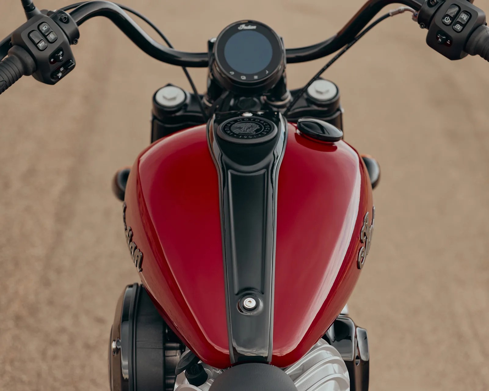 Glossy red motorcycle fuel tank with black center strip and handlebars on a sandy road.