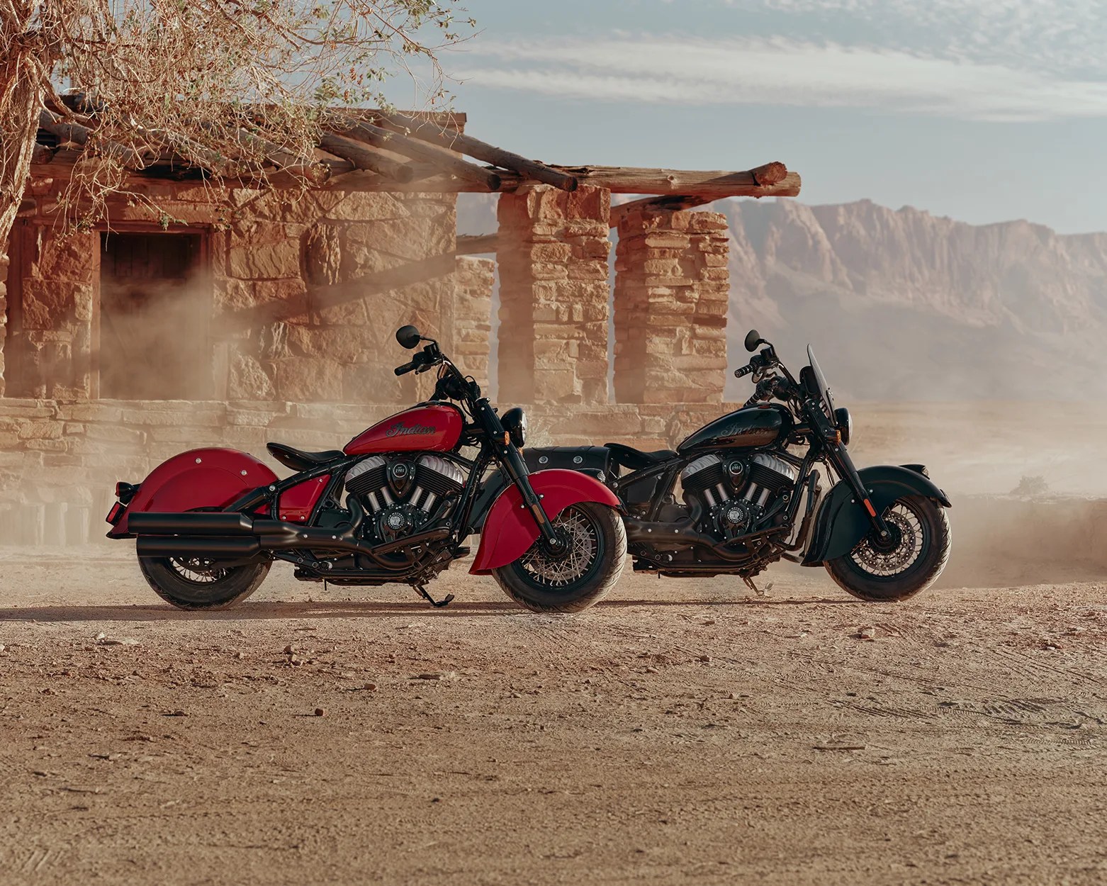Two Indian motorcycles, one red and one black, parked side by side on a dusty desert ground in front of a stone building.