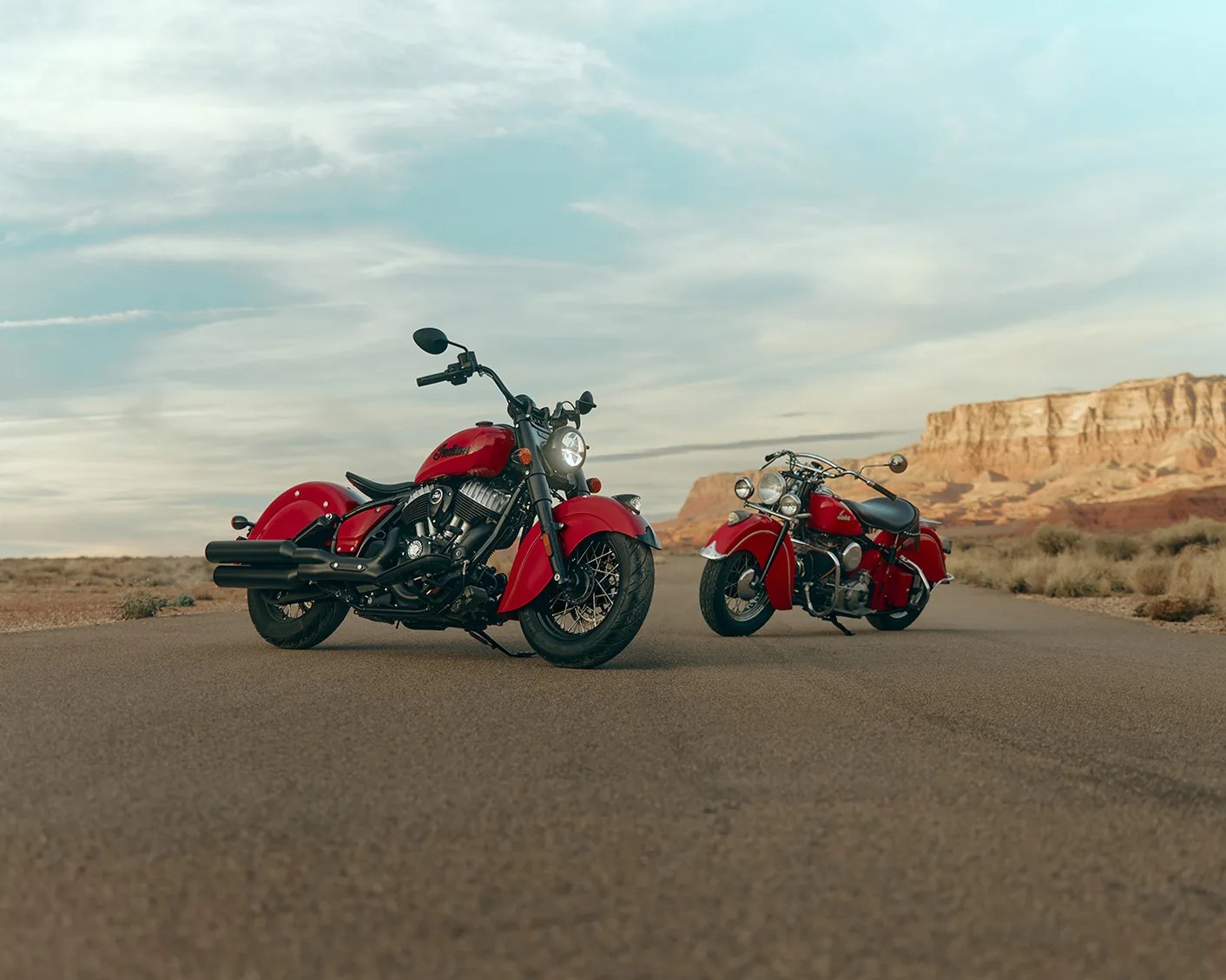 Two red Indian motorcycles parked on a desert road with rocky cliffs in the background.