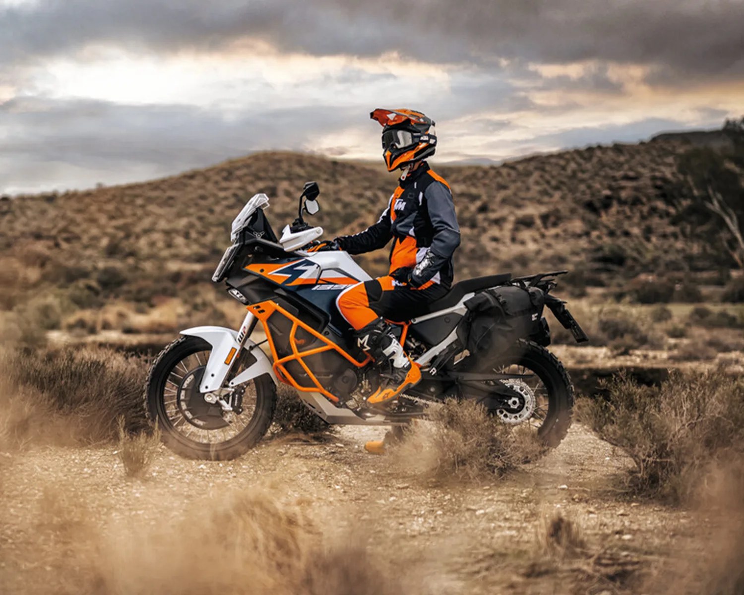 Person wearing orange and black riding gear sitting on a white and orange adventure motorcycle in a desert landscape.