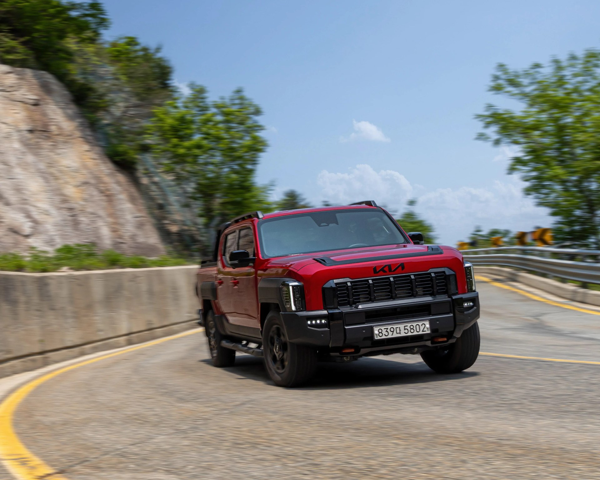 Red Kia pickup truck with black grille and accents driving on a curved mountain road.