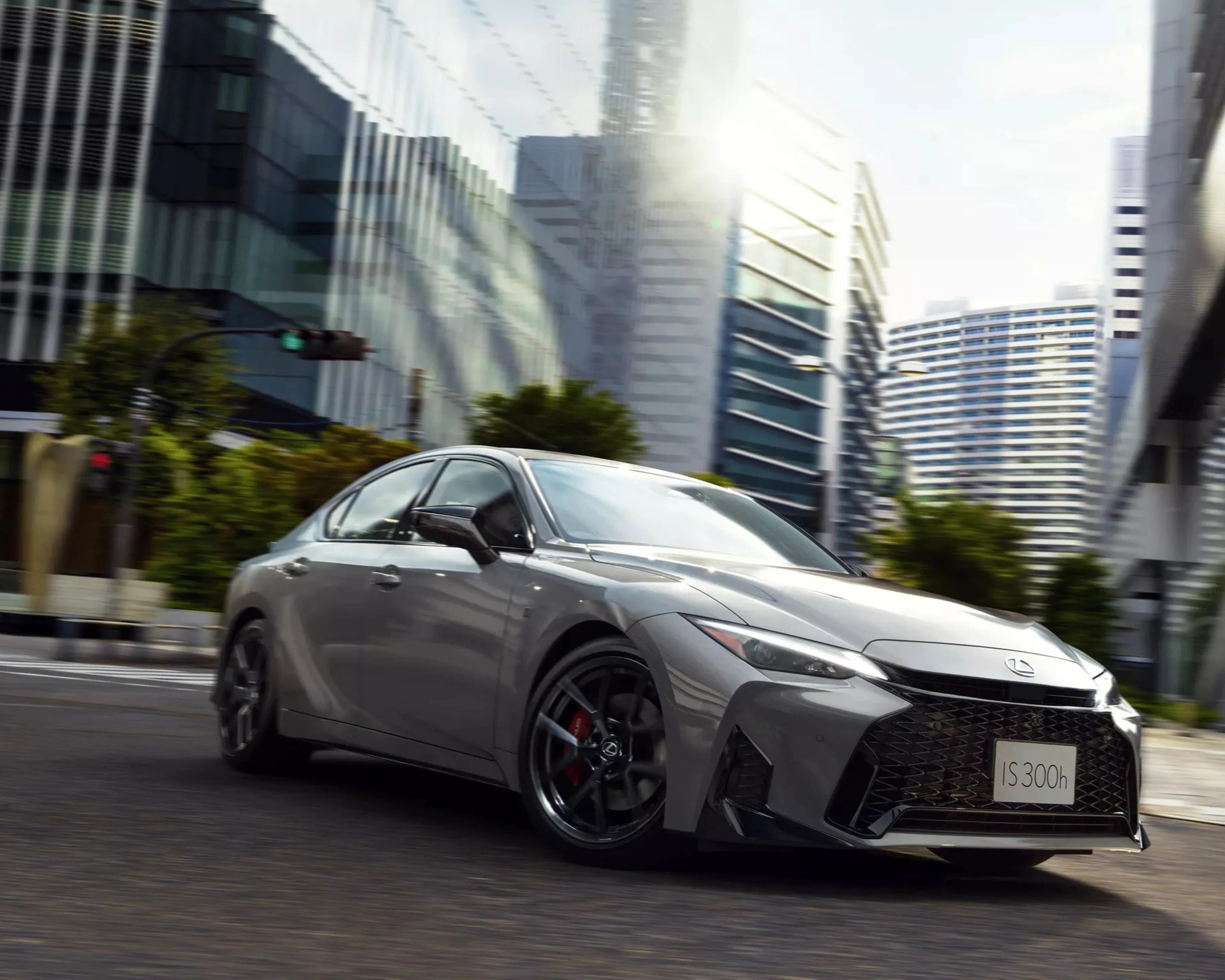 Gray Lexus IS 300h sedan with black wheels driving on a city street with modern buildings in the background.