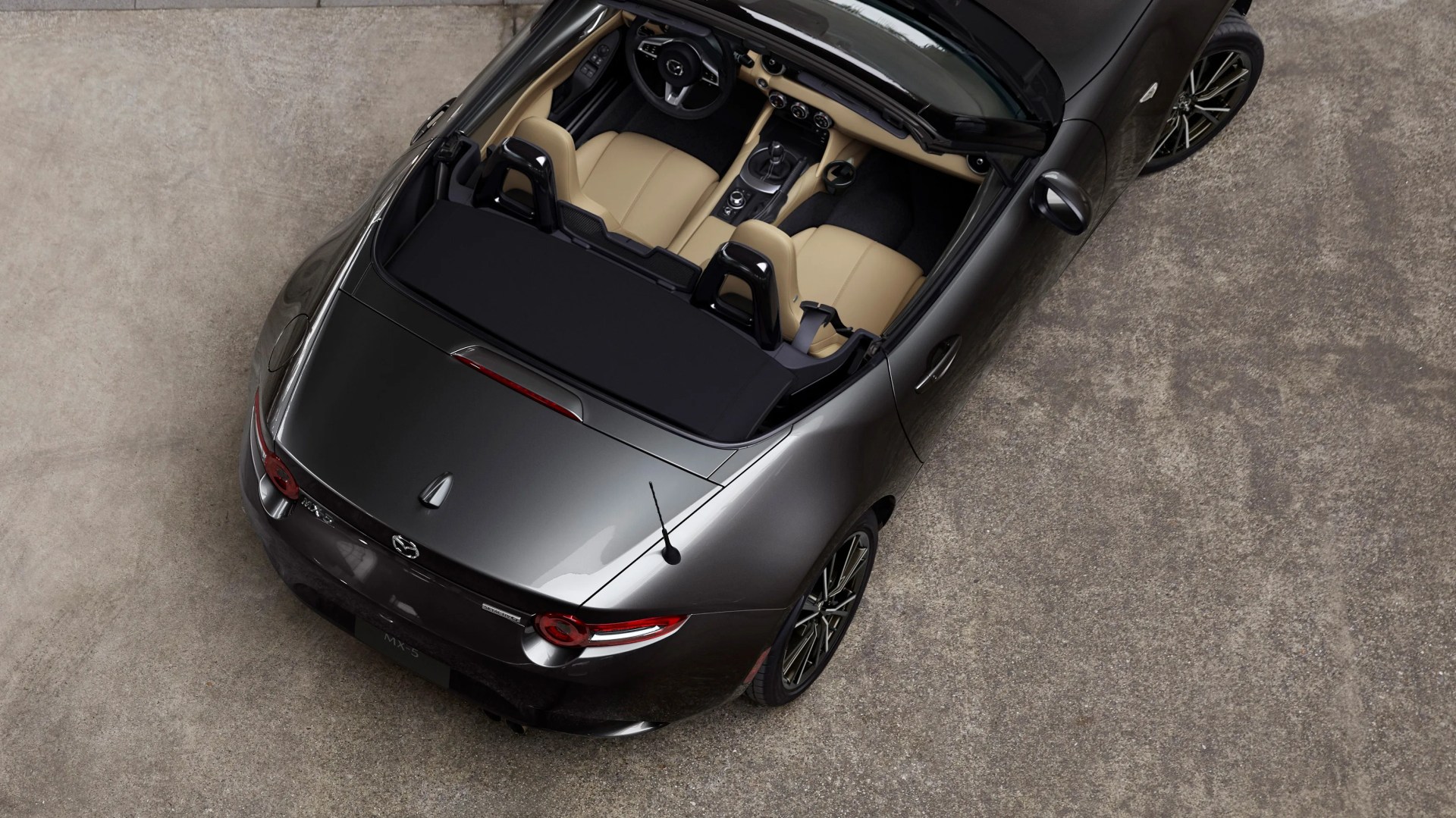 Dark gray Mazda MX-5 convertible with beige leather seats viewed from above on a concrete surface.