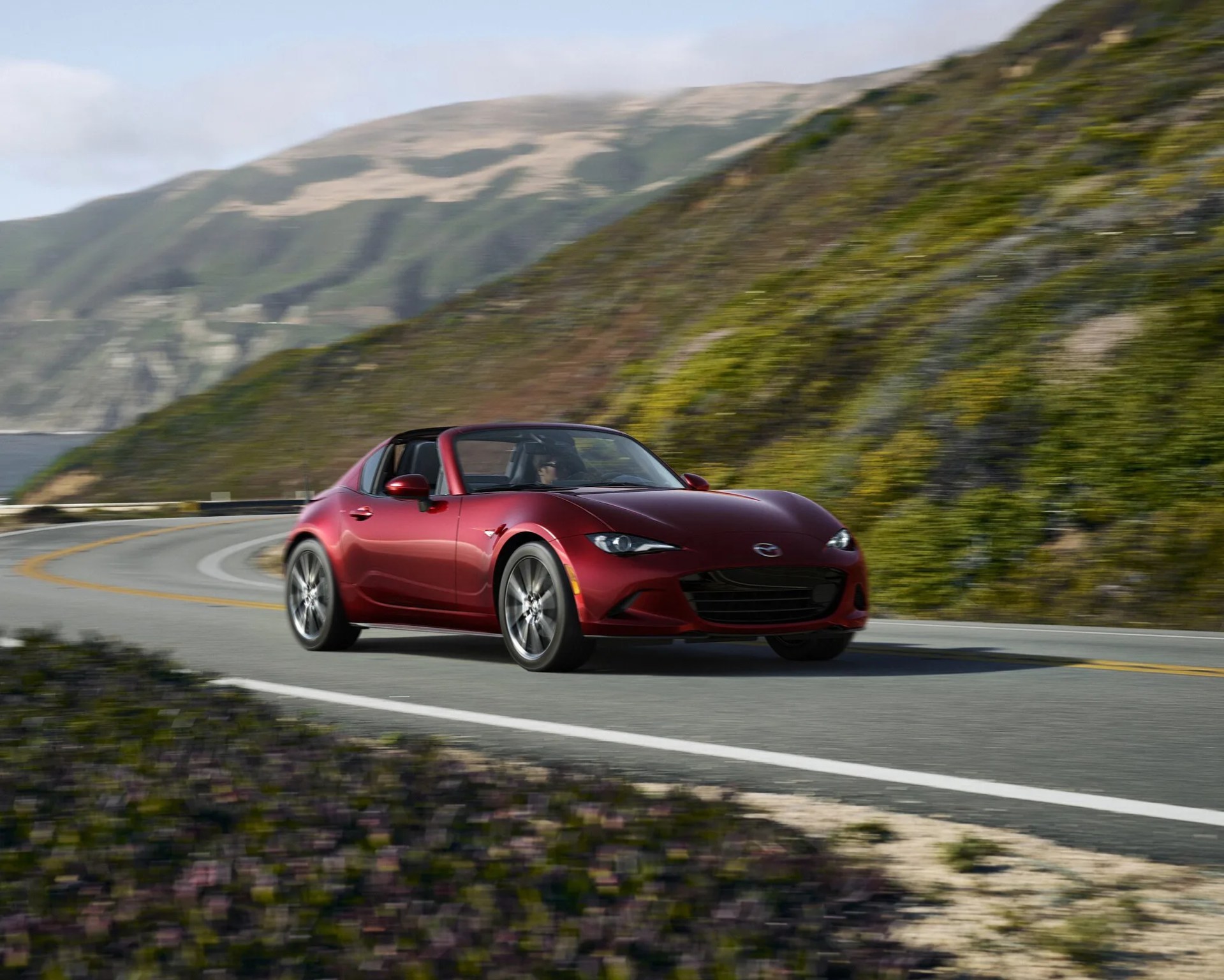 Red Mazda MX-5 Miata RF sports car driving on a winding mountain road with green hills in the background.