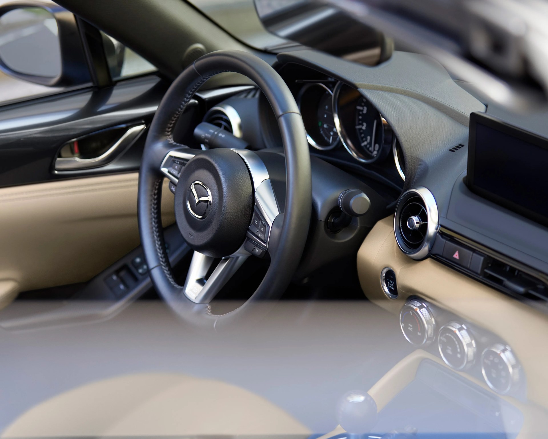 Black leather steering wheel with Mazda logo inside a beige and black car interior.
