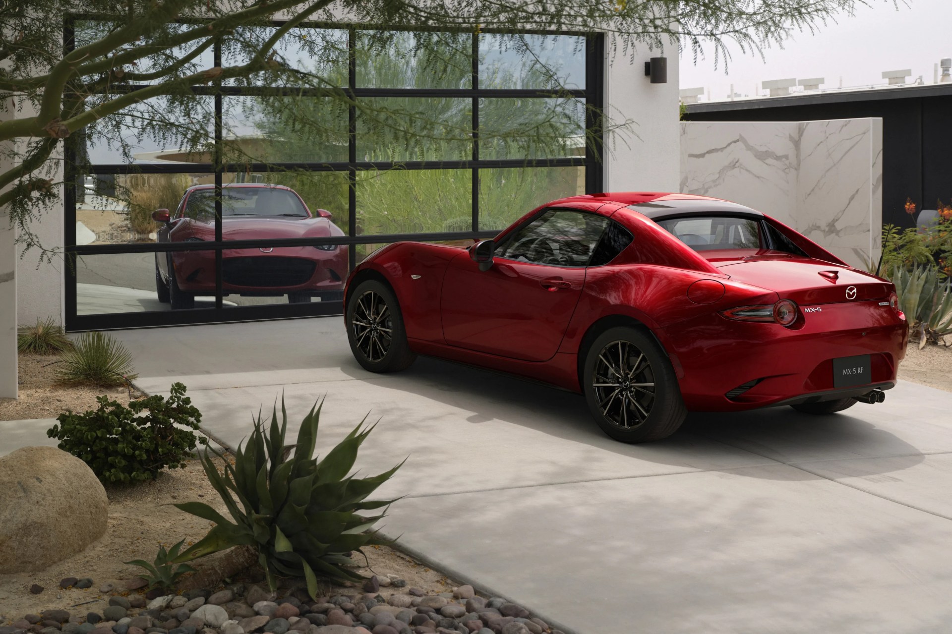 Red Mazda MX-5 RF sports car parked on a concrete driveway in front of a modern garage with glass doors.