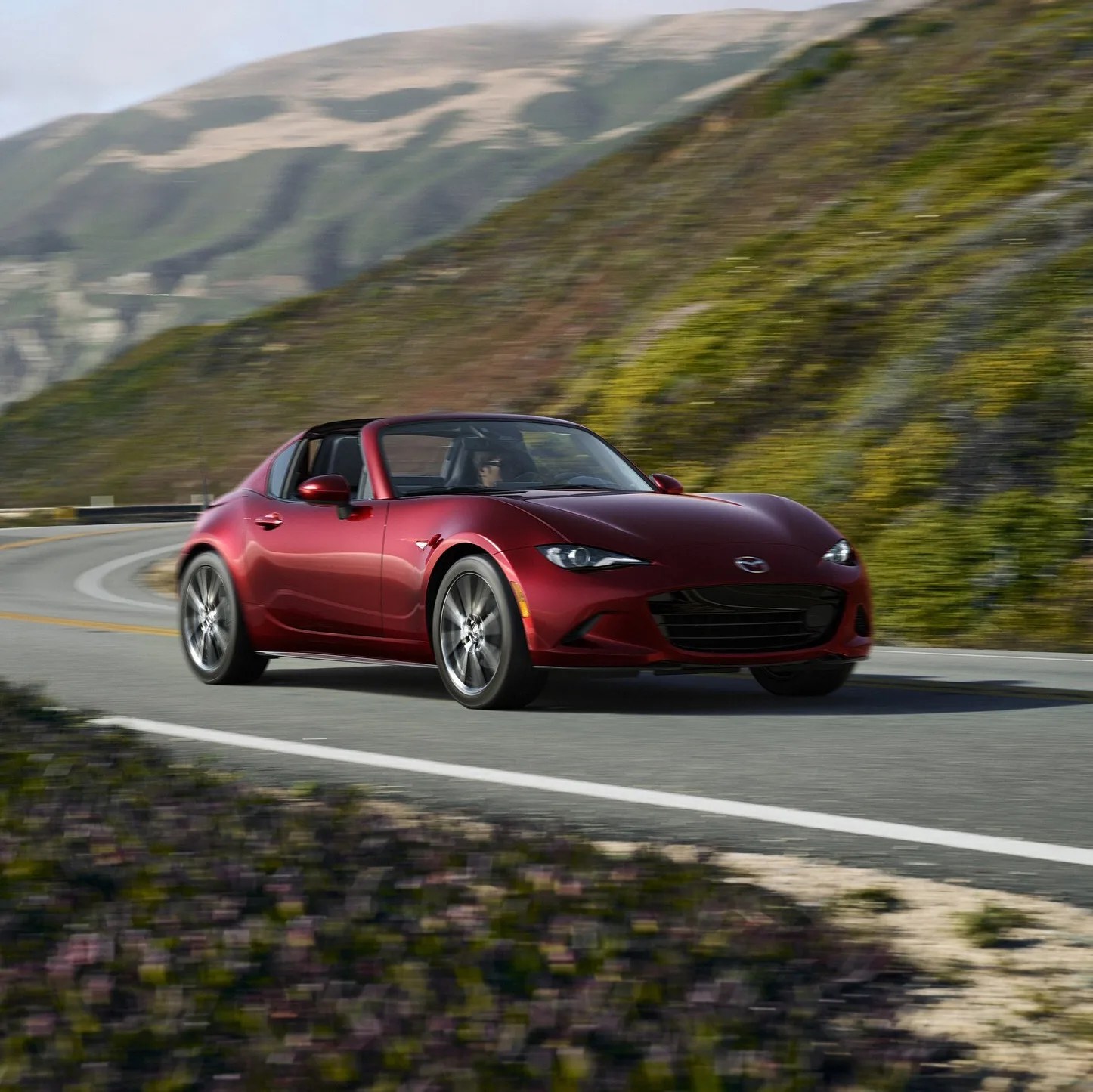 Red Mazda MX-5 Miata convertible driving on a winding mountain road with green hills in the background.