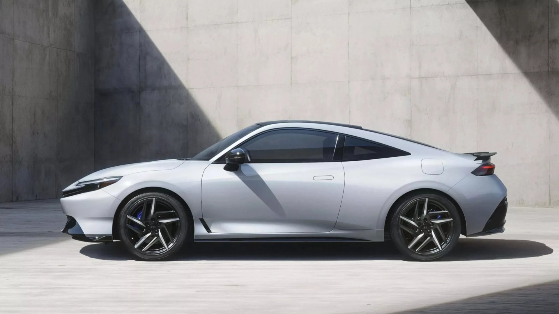 Silver two-door sports coupe with black alloy wheels parked in a concrete space with angular shadows.