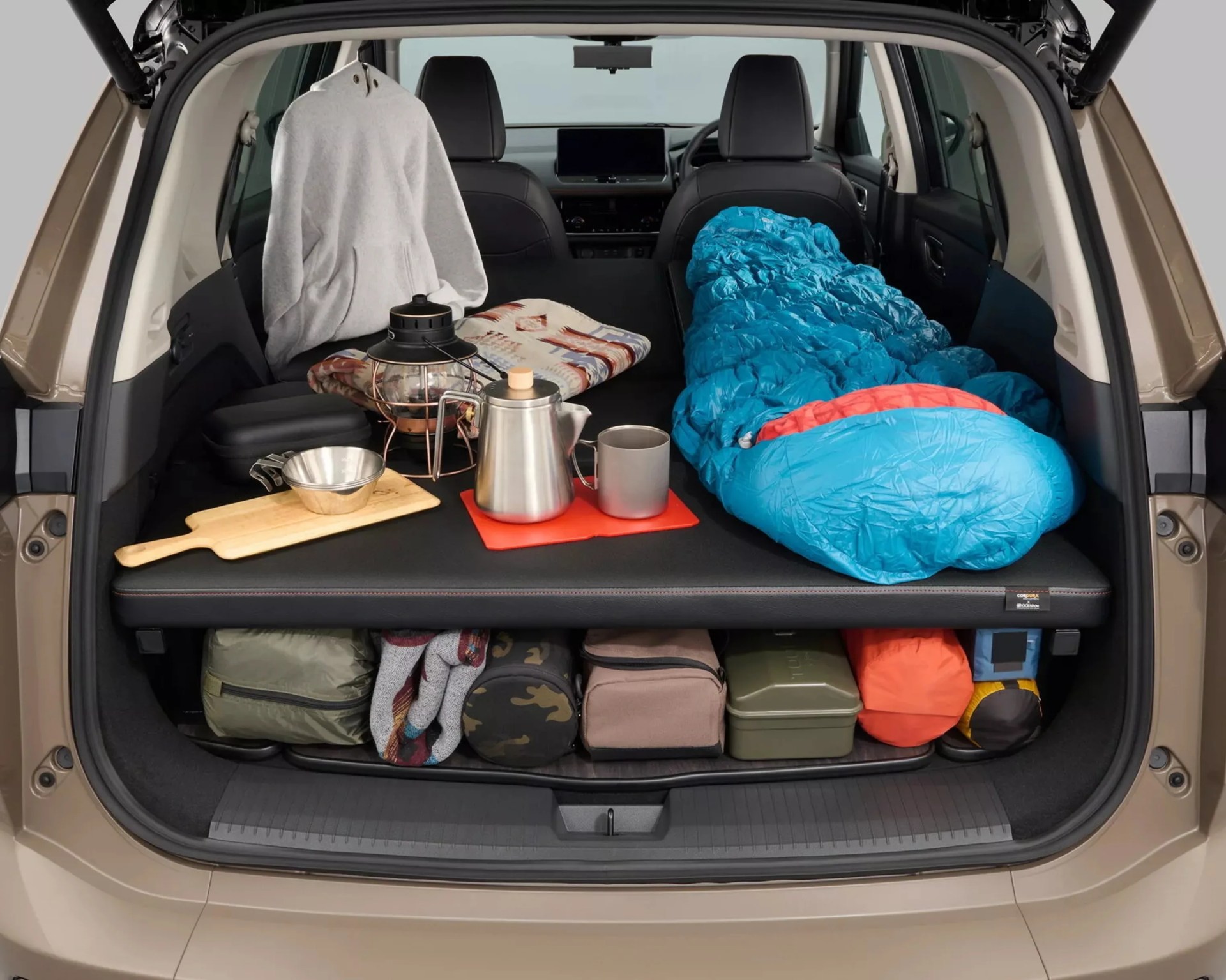 Car trunk set up for camping with a blue sleeping bag, gray hoodie, lantern, kettle, mug, and organized gear underneath a black platform.