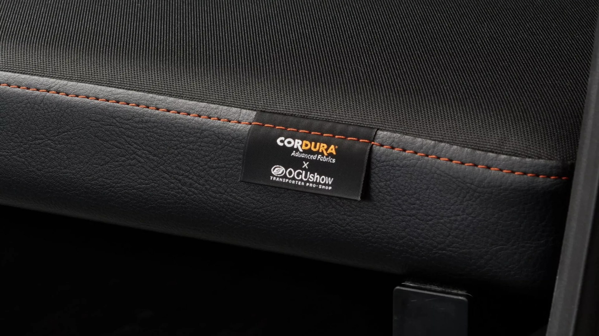 Close-up of black fabric and leather seat with orange stitching and a Cordura x OGushow label.