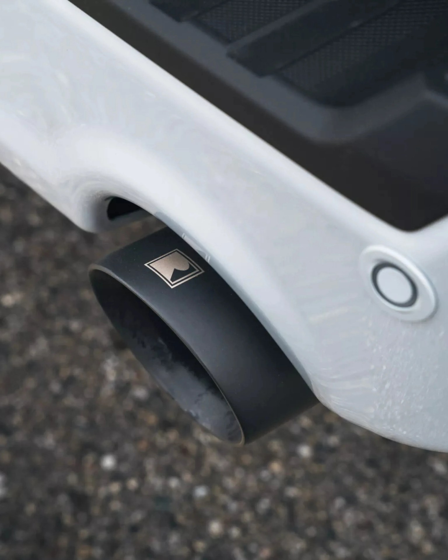 Black matte exhaust tip with a silver logo attached to a white vehicle bumper.